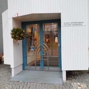 Valkoisen museorakennuksen ulko-ovi. Oven edustalla on metalliritilä. Ovessa näkyy invamerkki ja oven edessä roikkuu valaistu havupallo.