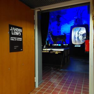 Hämärän näyttelytilan oviaukko. taustalla näkyy peura ja sinistä valoa.