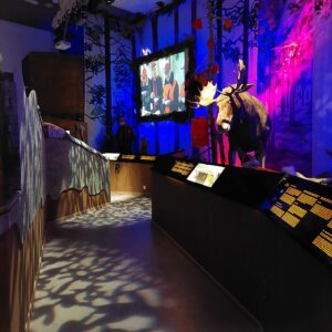 Hämärästi valaistu näyttelytila, jossa on kalteva kulkuliuska. Taustalla näkyy videoprojisointi sekä täytetty uroshirvi.