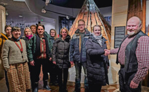 Iloisesti hymyileviä aikuisia museon näyttelytilassa. Kaikki katsovat kameraan ja oikealla nainen ja mies kättelevät.